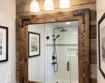 May include: A rustic bathroom mirror with a dark wood frame and black metal corner brackets. The mirror is mounted on a wooden plank wall, with a three-light fixture above. The reflection shows a shower and sink.