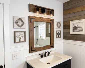 May include: Bathroom vanity with a large, rectangular mirror in a dark wood frame. Above the mirror is a black light fixture with three exposed bulbs. Decorative wall art and a white sink complete the rustic, farmhouse-style design.