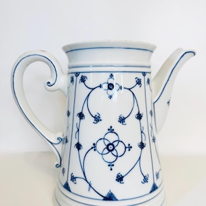 Puede incluir: Jarra de cerámica blanca con un diseño floral azul. La jarra tiene un asa curva y un pico. El patrón azul es simétrico y cubre el cuerpo. Probablemente se utiliza para servir bebidas.
