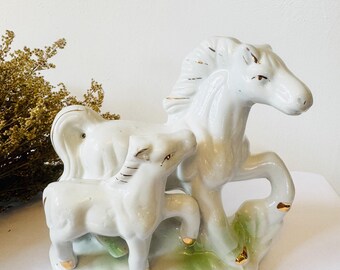 Puede incluir: Figuras de porcelana blanca de un caballo y un potro. El caballo está de pie sobre sus patas traseras con la cabeza girada hacia un lado. El potro está de pie junto al caballo y lo mira. Ambas figuras tienen detalles dorados.
