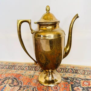 May include: A vintage brass coffee pot with a lid and a curved spout. The pot has a handle and sits on a pedestal base. The brass has a warm, golden hue, and the pot appears to be in good condition. The background is a patterned rug.
