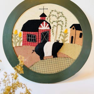 Puede incluir: Un adorno de pared redondo y enmarcado con una escena de granja. La obra representa un granero rojo con techo negro, un cerdo blanco y negro y una casita. El marco es de color verde oscuro.