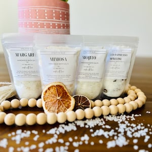 May include: Four small white bags of bath salts with labels that read "Margari", "Mimosa", "Mojito", and "Ed Espresso Martini". The bags are arranged in a row on a wooden surface with a string of wooden beads and dried citrus slices in front of them.