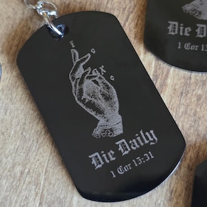 May include: A black metal dog tag keychain featuring a silver-toned laser-engraved image of a hand making a blessing gesture. The text "Die Daily" and "1 Cor 15:31" is engraved in a gothic font below the hand, with "IC XC" symbols above.