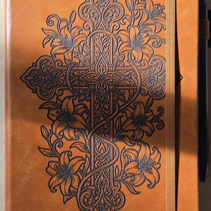 May include: A tan notebook with a dark brown embossed design on the front cover featuring a central cross intertwined with lily flowers and intricate scrollwork. The notebook includes a vertical elastic closure band and a black pen attached to the side.