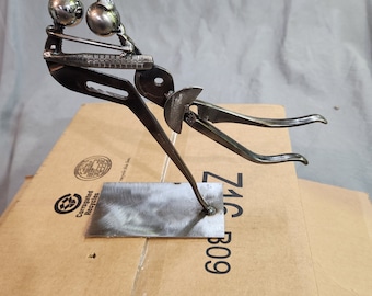 May include: A metal sculpture of a person holding two balls. The sculpture is made of recycled metal and is mounted on a metal base. The base is sitting on a cardboard box with the text "ZAC-B09" printed on it.