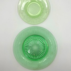 May include: Two green glass plates. The top plate is a lighter shade of green with a floral pattern around the rim. The bottom plate is a darker green with a textured surface and a ridged centre.