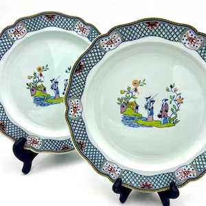May include: Two white ceramic plates with scalloped edges. Each plate features a central scene of figures near a body of water, surrounded by floral and geometric patterns in blue, red, and yellow. The plates are displayed on black stands.