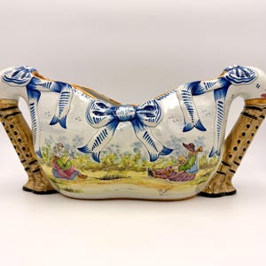 May include: A white ceramic decorative bowl with blue ribbon accents and handles. The bowl features a pastoral scene with figures playing instruments. The handles are brown with black accents.