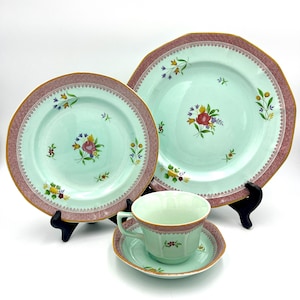 May include: A set of vintage porcelain tableware featuring a light green background with floral designs. The set includes plates, a teacup, and a saucer, all with a decorative burgundy and gold trim. The plates are displayed on black stands.