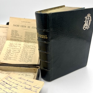 May include: A black leather-bound book with the word "MISSEL" in gold lettering. The book has a silver emblem and is next to open pages of text and handwritten notes. The book's pages have gold edges.