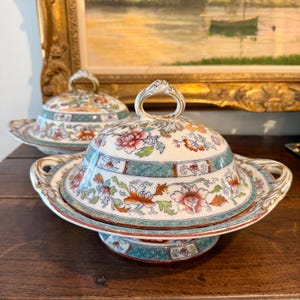 May include: A decorative, antique butter dish with a lid, featuring a floral design in shades of red, orange, and green on a cream background. The dish has a pedestal base and handles, with a ring-shaped handle on the lid. The dish is placed on a wooden surface.