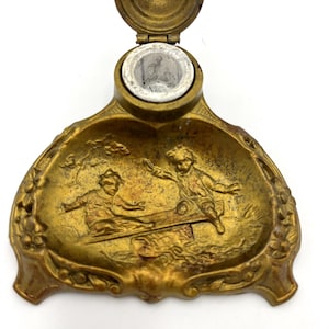 May include: Antique brass inkwell with a hinged lid and a detailed relief scene of children playing on a seesaw. The inkwell has a decorative, ornate design with floral accents around the edges. The inkwell is a vintage item.