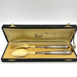 May include: A vintage silverware set in a black case. The set includes a fork, spoon, and serving spoon with ivory-colored handles and silver-toned metal accents. The case has a yellow interior and a gold clasp.