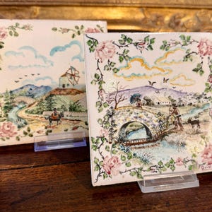 May include: Two square ceramic tiles with hand-painted landscape scenes. One depicts a windmill and a person on a horse, the other a stone bridge and figures. Both are framed with pink roses and green foliage.