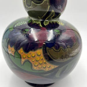 May include: A decorative ceramic vase with a bulbous base and a smaller top section. The vase features a glossy finish and is adorned with a colorful floral pattern in shades of purple, green, orange, and gold. The vase is approximately 8 inches tall.