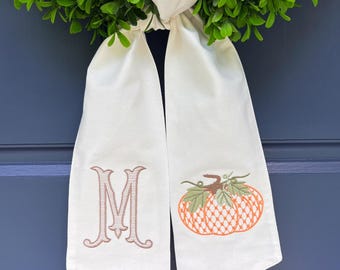 May include: A white fabric door hanger with an embroidered initial "M" and a pumpkin design. The hanger is adorned with a green wreath and the text "magnolia momma embroidery". The background is a blue door.