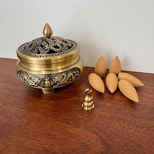 May include: A gold metal incense burner with a dragon design and a lid with a tree design. The burner is on a wooden surface with six brown incense cones.