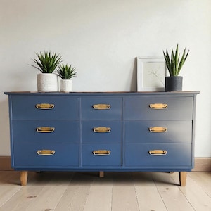 Op de afbeelding: Een blauwe dressoir met negen lades en goudkleurige handgrepen. De dressoir heeft een houten bovenkant en staat op vier houten poten. Twee potplanten en een ingelijste foto staan erop.