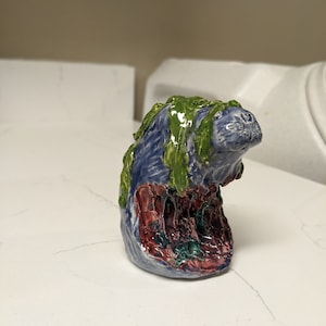Peut inclure: Une sculpture en céramique d'une créature avec un corps bleu, de la mousse verte et des détails rouges et violets. La créature a une tête ronde et une large bouche.