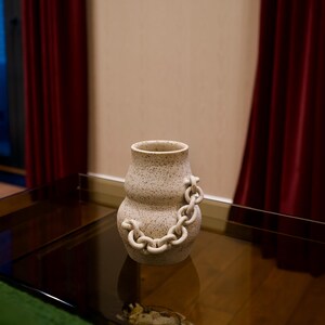 Könnte beinhalten: Eine gesprenkelte beige Keramikvase mit einem Kettenmuster. Die Vase hat eine abgerundete Form mit einem schmalen Hals und einer kettenartigen Verzierung, die sich um ihren Körper wickelt. Die Vase steht auf einem Glastisch.