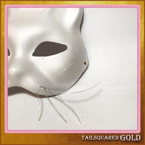 May include: A white cat mask with eye cutouts and wire whiskers. The mask is made of a smooth, reflective material. The text "TAILSQUARED GOLD" is at the bottom of the image.