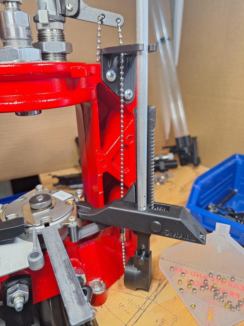 May include: A close-up of a reloading press with a black metal clamp attached to a silver rod. The clamp has a black lever and the text "SMALL" printed on it. A silver chain is attached to the clamp and the press.