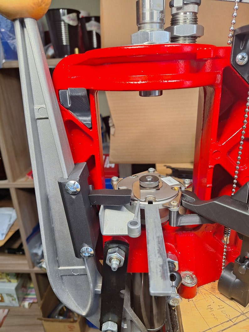 May include: A red reloading press with a silver lever and a black handle. The press has a silver metal plate with a small black lever on the right side.