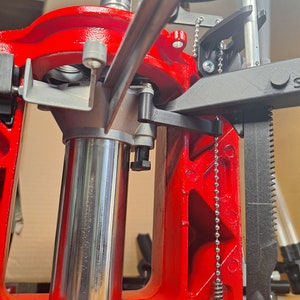 May include: Close-up of a red reloading press with a silver metal cylinder and a black lever. A chain with a spring-loaded hook hangs from the side of the press.