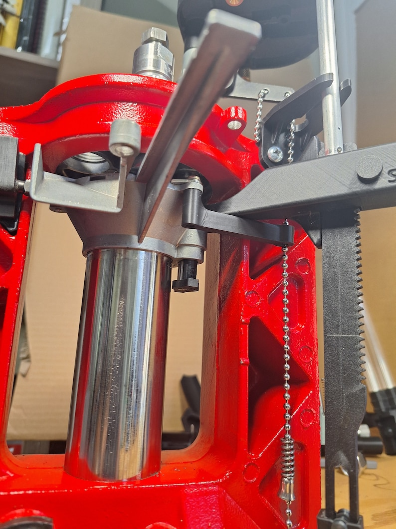 May include: Close-up of a red reloading press with a silver metal cylinder and a black lever. A chain with a spring-loaded hook hangs from the side of the press.