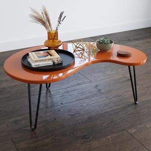 Peut inclure: Une table basse en forme de rein avec une surface orange brillante et trois pieds en épingle en métal noir. Sur la table se trouvent un plateau circulaire noir contenant deux livres intitulés Mid-Century Form et Eames, un vase en verre ambré avec des tiges séchées, un objet géométrique en fil doré, une petite plante succulente et trois sous-verres marron.