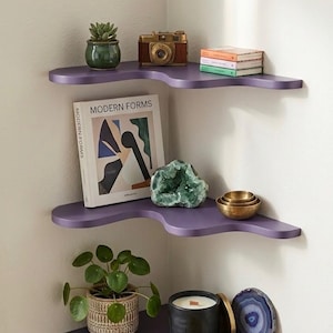 Puede incluir: Estanter&iacute;a esquinera de tres niveles en color p&uacute;rpura, con dise&ntilde;o ondulado. Las estanter&iacute;as exhiben una colecci&oacute;n de objetos: una planta, una c&aacute;mara vintage, libros, una geoda, una vela y una piedra decorativa.