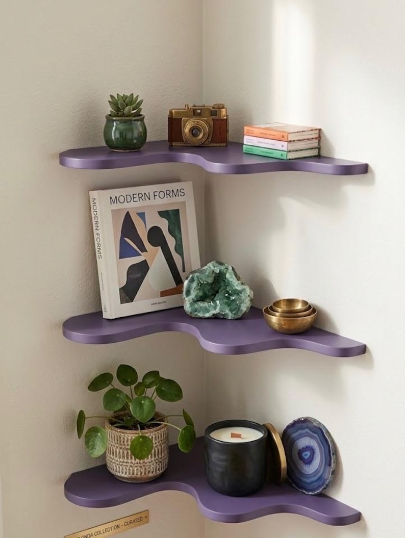 Puede incluir: Estanter&iacute;a esquinera de tres niveles en color p&uacute;rpura, con dise&ntilde;o ondulado. Las estanter&iacute;as exhiben una colecci&oacute;n de objetos: una planta, una c&aacute;mara vintage, libros, una geoda, una vela y una piedra decorativa.