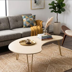 Può includere: Un tavolino a forma di rene beige chiaro con gambe a forcina dorate. Un vassoio dorato con tazza e piattino è appoggiato sul tavolo. Sullo sfondo un divano grigio, un cuscino fantasia e una coperta gialla. Sotto il tavolo un tappeto beige con un motivo geometrico.