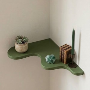 Puede incluir: Una estanter&iacute;a de esquina verde con un dise&ntilde;o &uacute;nico y fluido. Contiene una peque&ntilde;a planta suculenta en maceta, un cristal verde, una pila de libros antiguos y una vela verde en un portavelas de lat&oacute;n. La estanter&iacute;a est&aacute; montada en una pared de color neutro.