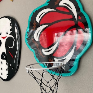 May include: A turquoise, black, white, and red basketball hoop with a black net. The basketball hoop is shaped like a cartoon basketball with a mouth and teeth.