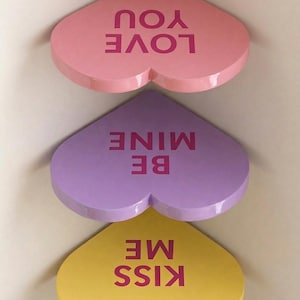 Op de afbeelding: Drie hartvormige planken in roze, paars en geel. De roze plank leest "LOVE YOU", de paarse plank "BE MINE" en de gele plank "KISS ME". Perfect voor Valentijnsdag of een romantisch cadeau.