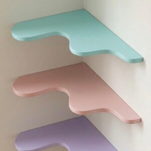 Puede incluir: Tres estanter&iacute;as de esquina en colores pastel: azul claro, rosa y lavanda. Cada estanter&iacute;a tiene un dise&ntilde;o curvo &uacute;nico en el borde frontal. Est&aacute;n montadas en una pared blanca, creando una est&eacute;tica moderna y minimalista.