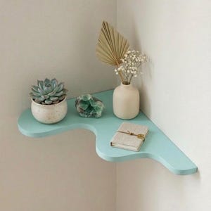 Puede incluir: Una estanter&iacute;a de esquina azul claro con una suculenta en una maceta blanca, un cristal verde, un peque&ntilde;o jarr&oacute;n blanco con flores secas y un peque&ntilde;o libro. La estanter&iacute;a est&aacute; montada en una pared blanca.
