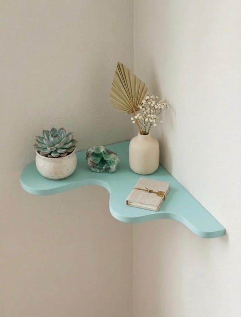 Puede incluir: Una estanter&iacute;a de esquina azul claro con una suculenta en una maceta blanca, un cristal verde, un peque&ntilde;o jarr&oacute;n blanco con flores secas y un peque&ntilde;o libro. La estanter&iacute;a est&aacute; montada en una pared blanca.