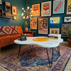 Peut inclure: Salon avec un canapé orange brûlé, une table basse blanche en forme d'œuf et un tapis vintage. Le mur est décoré d'œuvres encadrées et d'une enseigne au néon « EGGS & ART ».