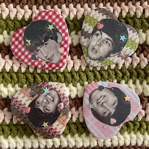 May include: Four heart-shaped pins featuring black and white portraits of The Beatles. Each pin has a unique background pattern including red gingham, floral, and striped designs, accented with small holographic star and heart-shaped stickers.