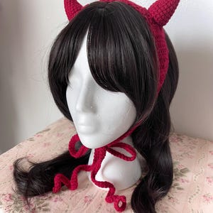 May include: A deep red crochet bonnet featuring two prominent, pointed devil-style horns on top. The headpiece is constructed with a textured yarn stitch and includes long, thin crocheted ties that fasten under the chin with a bow.