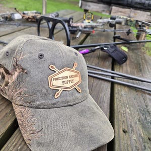 Op de afbeelding: Grijze baseballcap met een houten patch met de tekst "PRECISION ARCHERY SUPPLY". De cap heeft een camouflage-ontwerp en ligt op een houten tafel. Boogschietapparatuur is zichtbaar op de achtergrond.