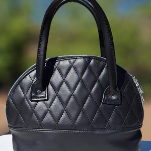 Könnte beinhalten: Schwarze, gesteppte Handtasche mit rundem Oberteil und stabilem Griff. Die Tasche hat ein Rautenmuster und einen silbernen Reißverschluss. Der Boden der Tasche ist einfarbig schwarz. Der Hintergrund ist verschwommen, mit grünen und blauen Farbtönen.