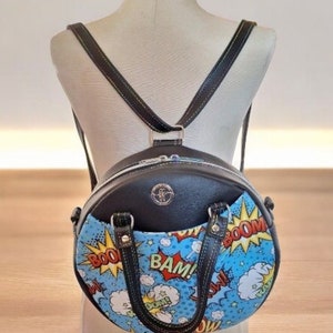 以下が含まれることがあります： 前面に青と白のコミック柄が施された、丸い黒革のバックパック。プリントには「BAM！」と「BOOM！」の文字が赤と黄色で書かれています。