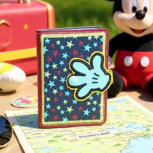 May include: A navy blue passport holder with a star pattern and a Mickey Mouse glove applique. The passport holder has a red border and yellow stitching. A Mickey Mouse plush toy is in the background.
