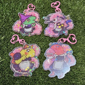 Könnte beinhalten: Vier bunte Schlüsselanhänger mit Comicfiguren, die als Dienstmädchen verkleidet sind. Jeder Schlüsselanhänger hat einen rosa herzförmigen Anhänger und einen rosa floralen Hintergrund. Die Figuren sind ein grünes Eidechsen, ein rosa-violettes Wesen, ein gelbes Wesen und ein rotes Wesen.