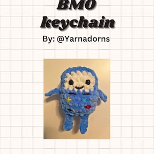 Op de afbeelding: Een handgemaakte blauwe en witte BMO sleutelhanger. De pluche sleutelhanger heeft een lachend gezicht, armen en benen. De tekst "BMO keychain" en "By: @Yarnadorns" staan erboven.