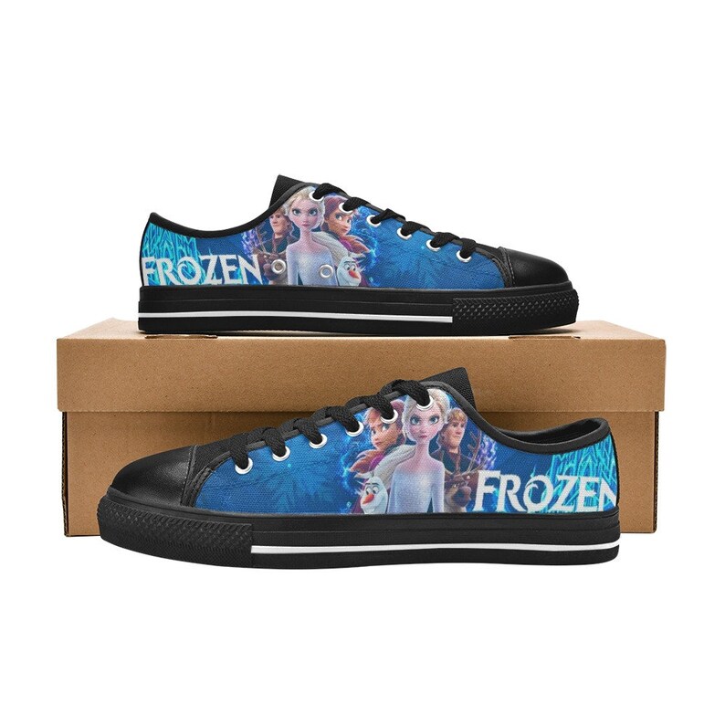 Puede incluir: Zapatillas bajas de lona negras con un estampado azul y blanco con personajes de la pel&iacute;cula Frozen. El estampado incluye a Elsa, Anna, Kristoff y Olaf. Las palabras "Frozen" est&aacute;n impresas en el lateral de cada zapato.